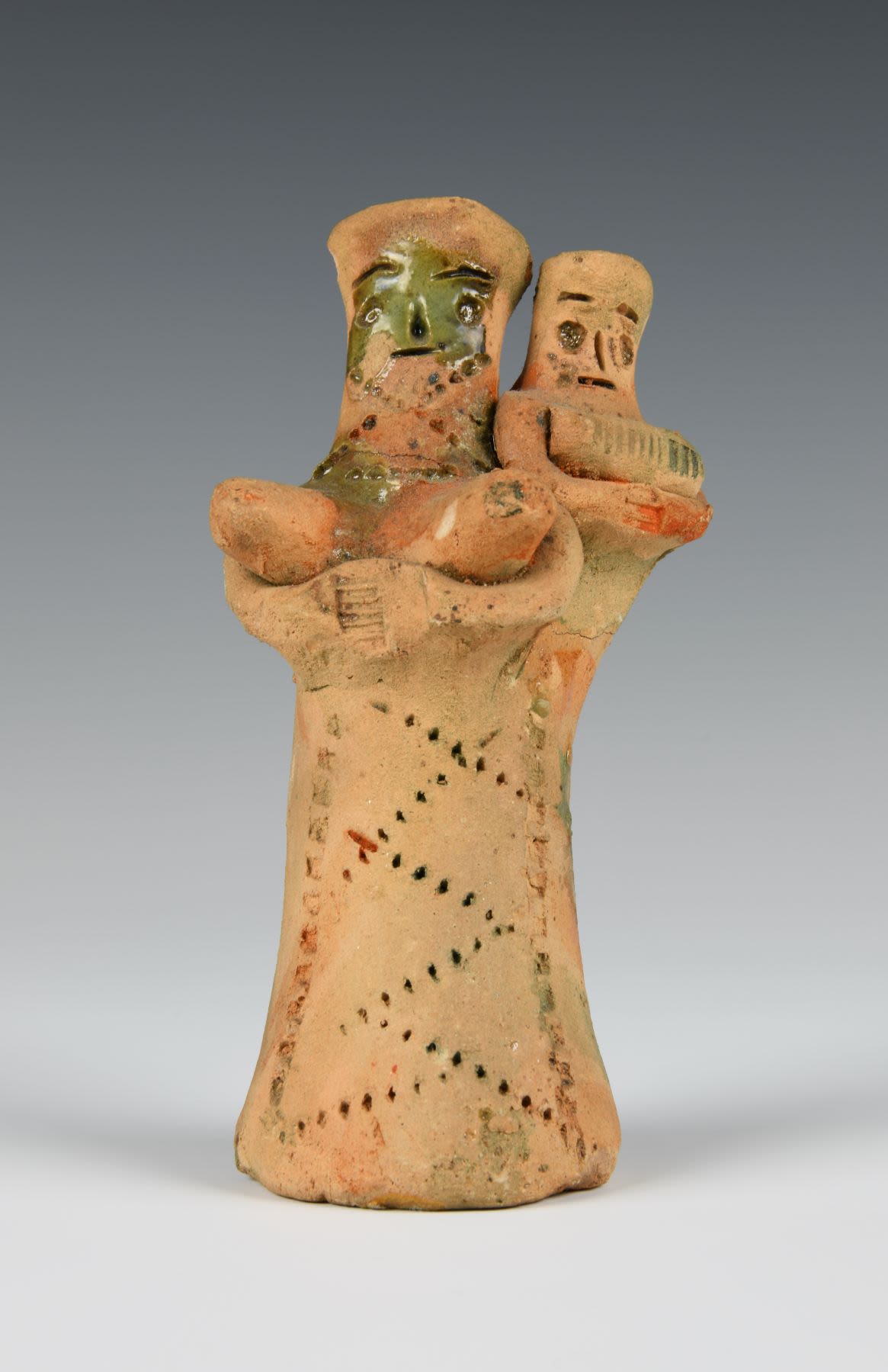 Moulded painted clay figure depicting a nurse with a child. The toy is unpainted with zigzag decoration on the woman's body and a faded stripe pattern in blue and red on both figures. Small flakes of green glaze cover some parts of the woman's face and right shoulder.