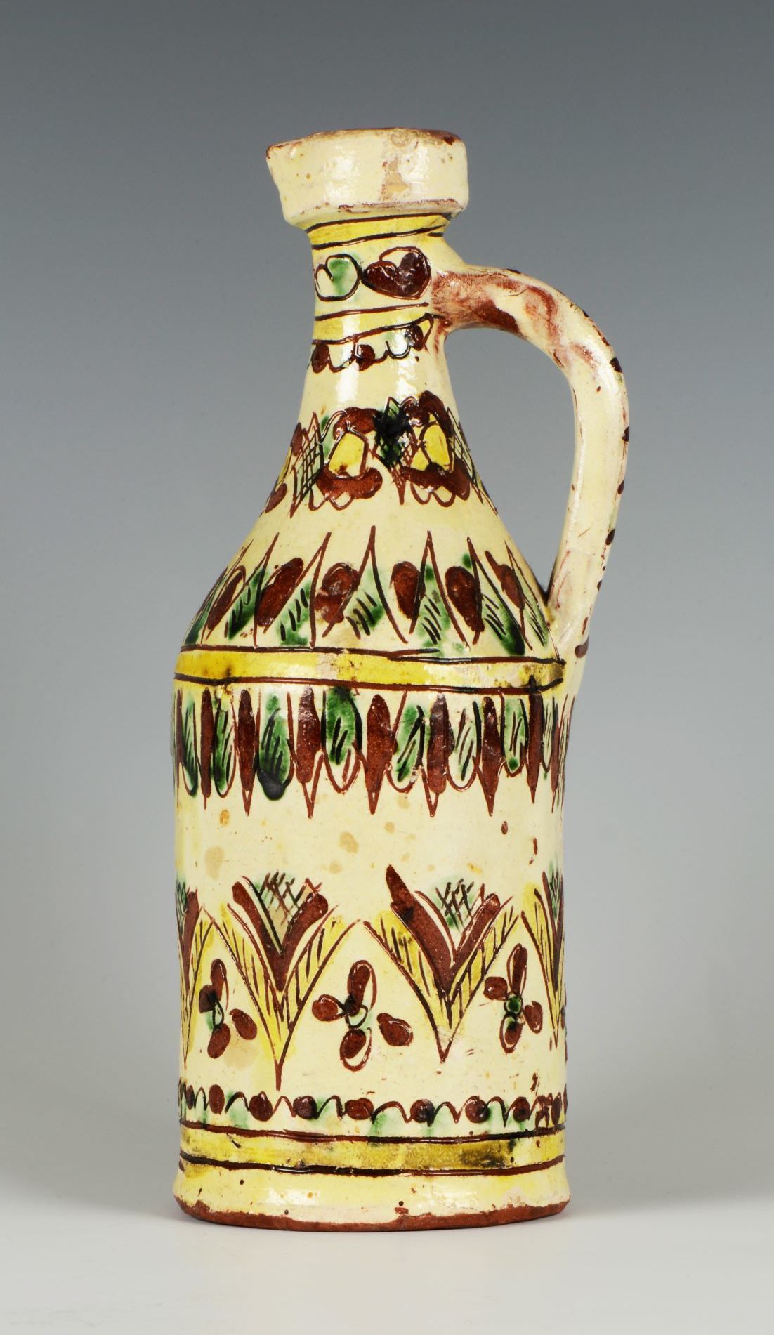 A ceramic glazed jug with a handle from one side. The jug has painted flowers, possibly leaves and geometric patterns painted in green, yellow and brown on a faded yellow background.