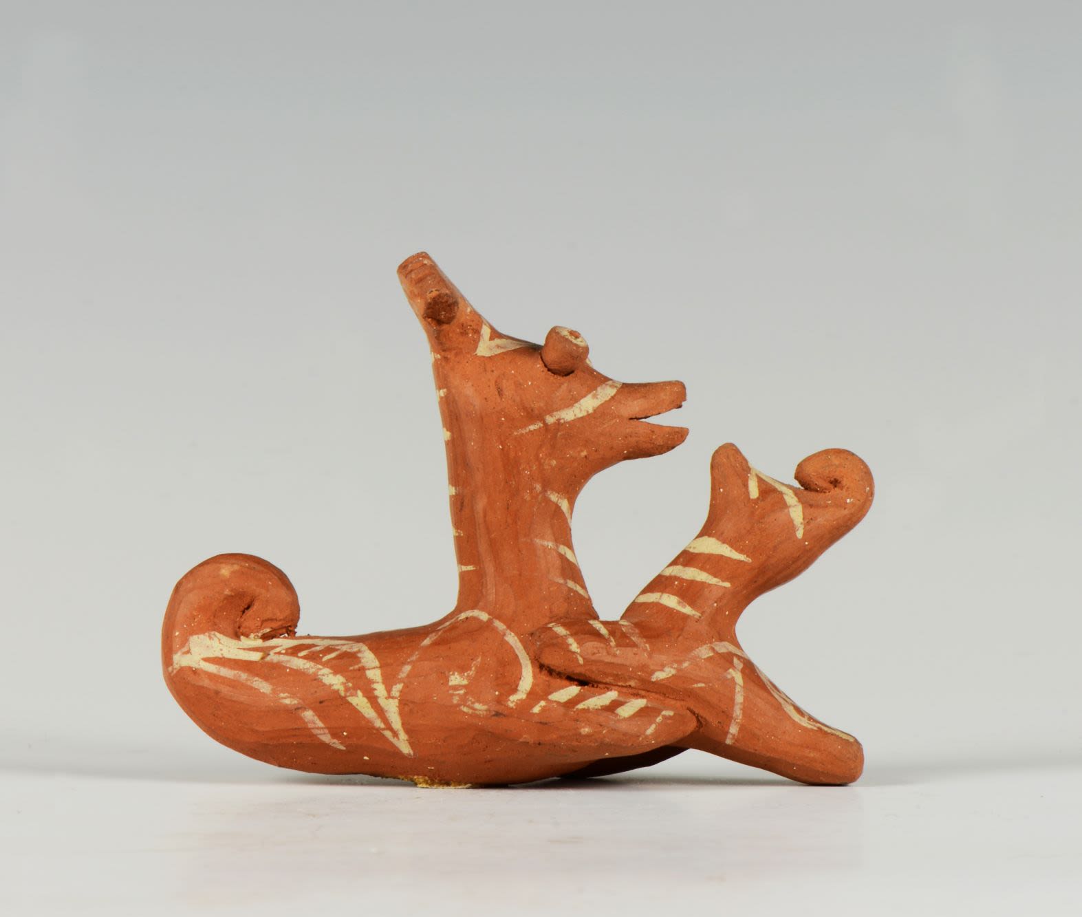 Clay figure of fantastical creatures facing each other. They are both unpainted but decorated with a white geometric motifs.