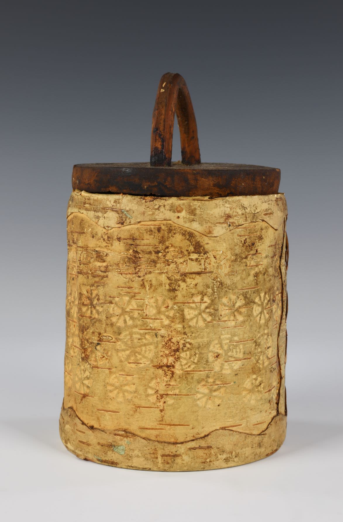 Birch bark container with lid. The top and the bottom of the container is decorated with a wavy rim. The exterior is decorated with a round pattern made of embossed triangles. At the base of the container is an indecipherable script in Russian.