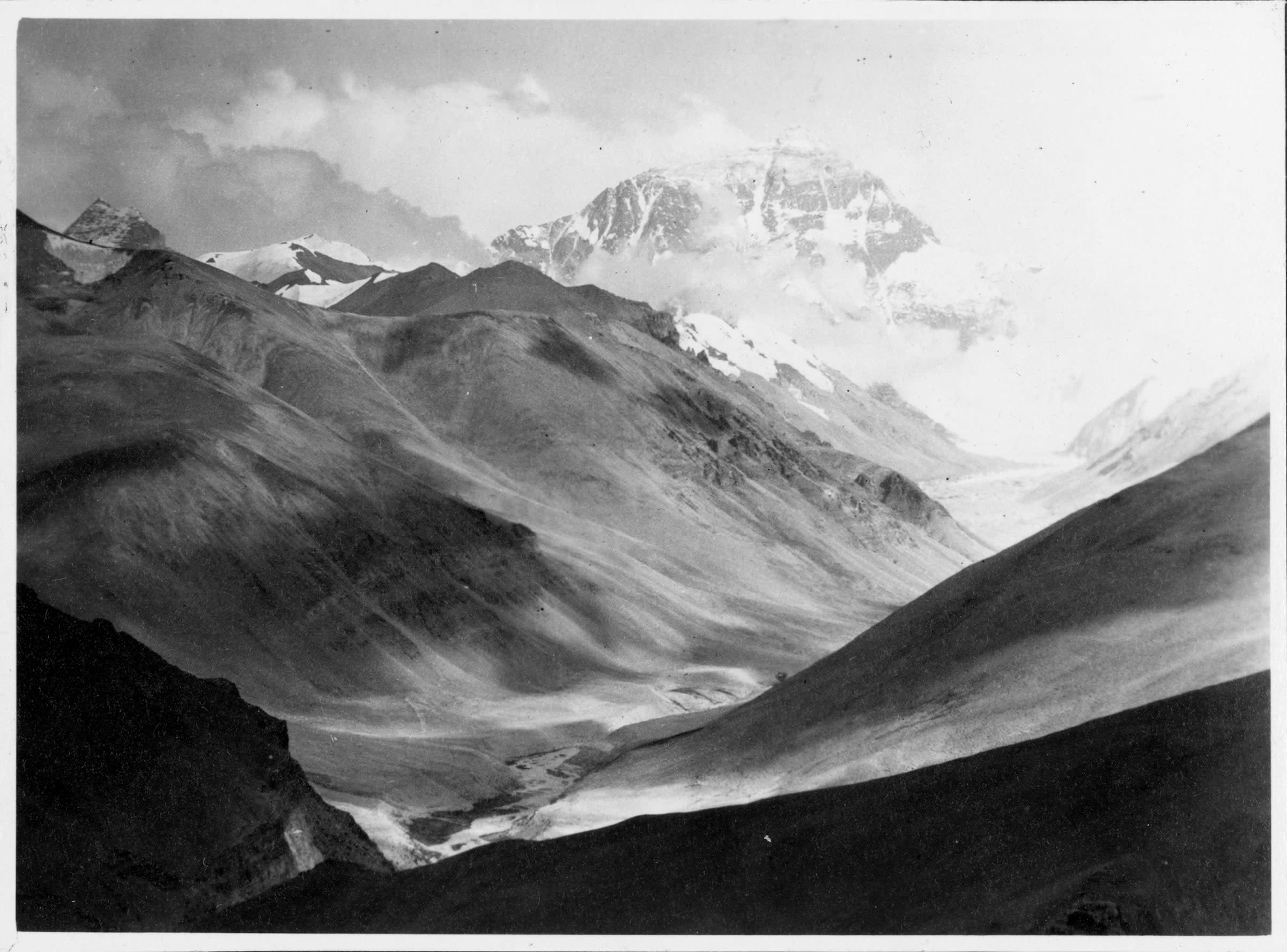 Black and white photo of a large mountain range. A tall snow covered mountain appears in the distance emerging from the clouds. 