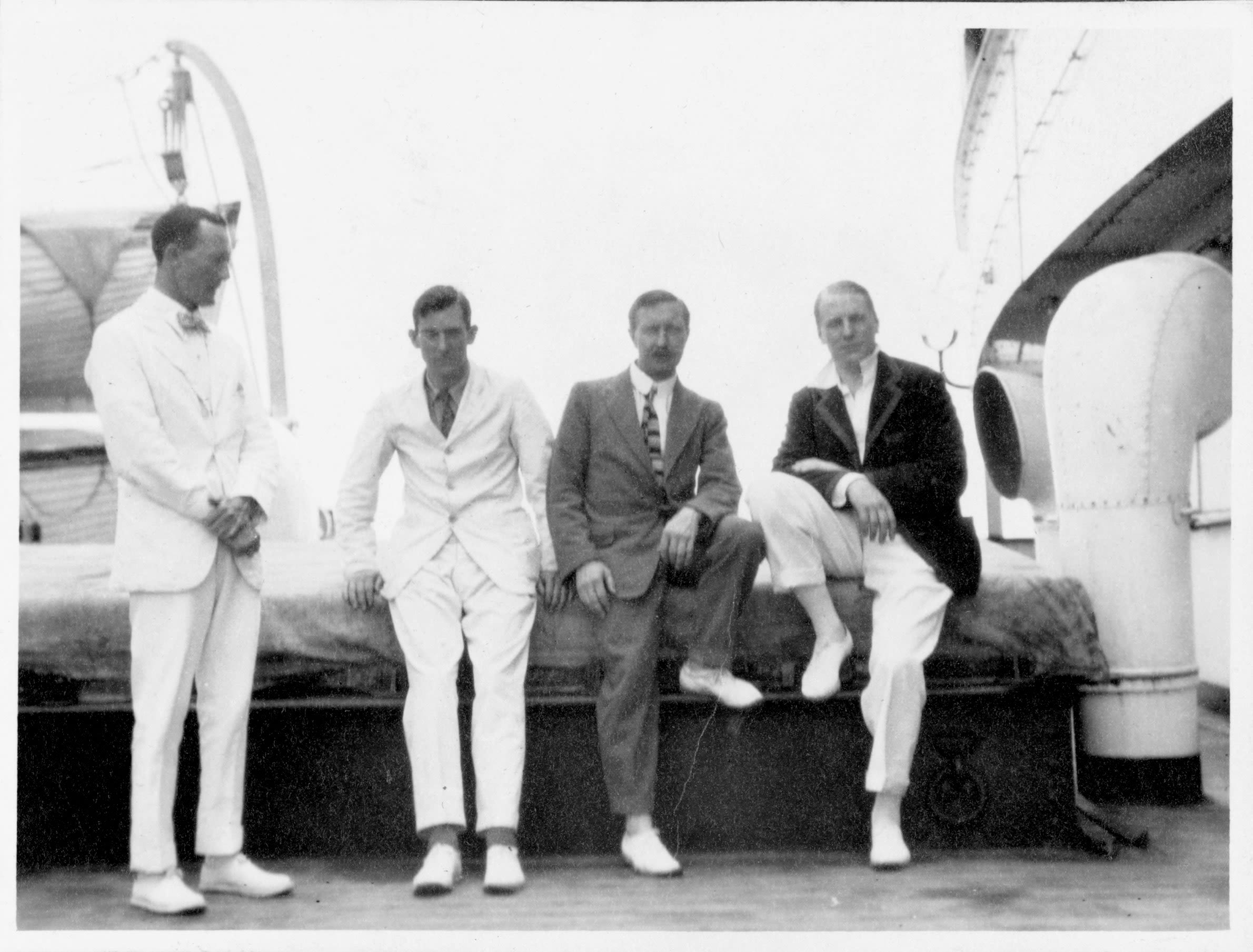 Black and white photo of four men posed for the camera on the deck of a boat. The men are smartly dressed in suits and ties, including bright white shoes and a plush velvet jacket.
