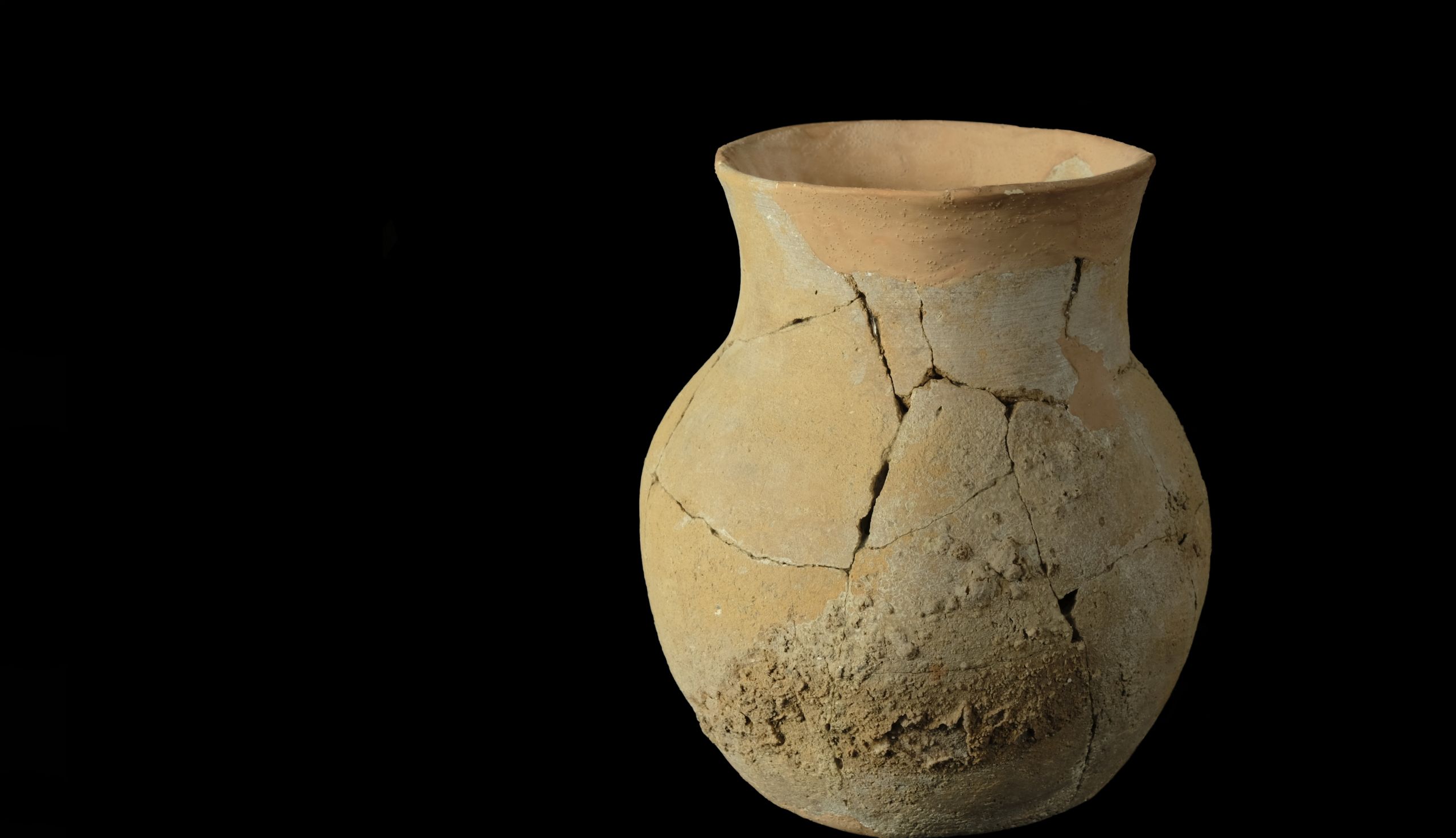 A tall, wide-rimmed pot on a black background. The pot is reconstructed with orange plaster from many dirt-encrusted sherds 