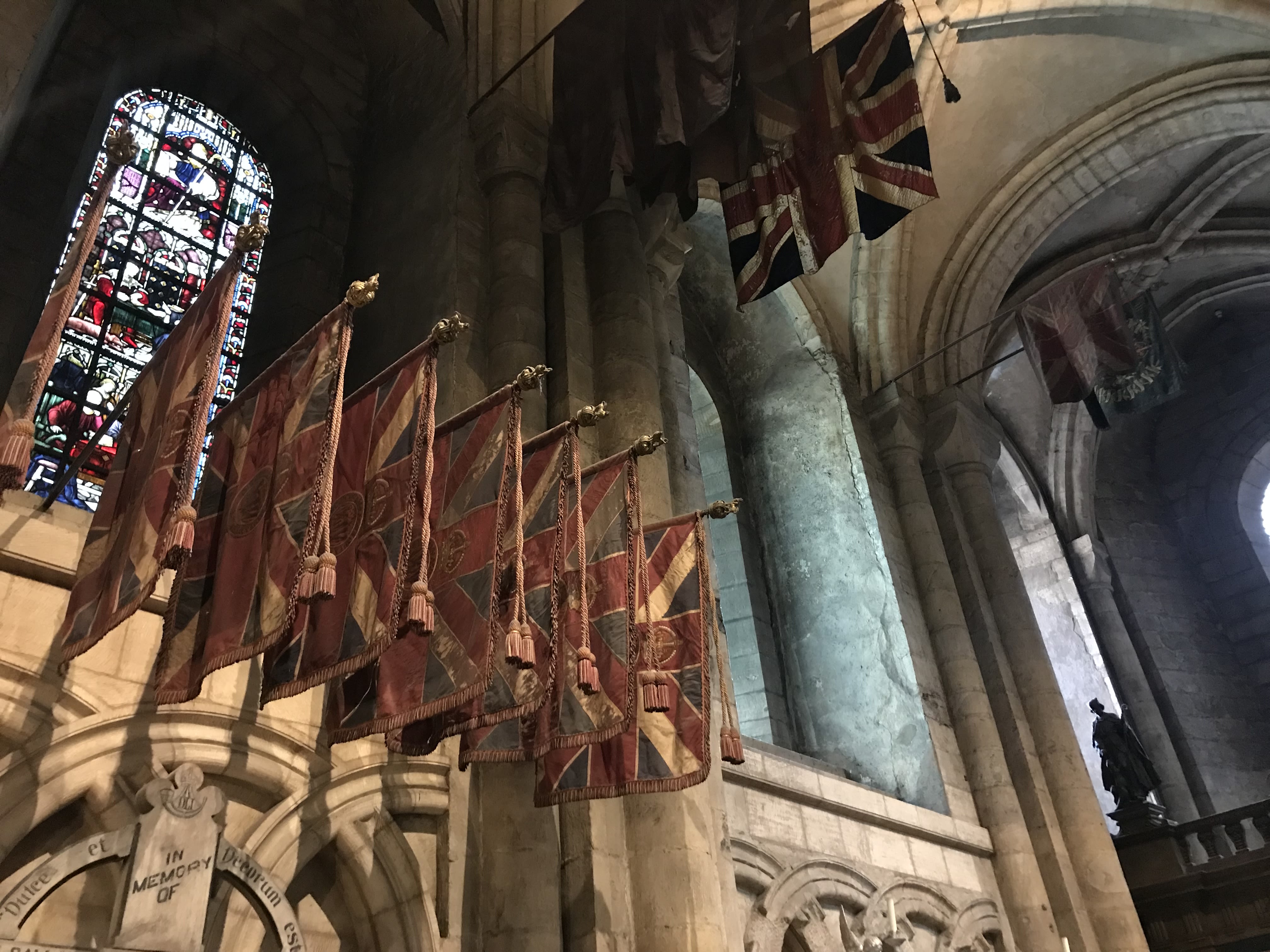 Photo of the DLI Chapel at Durham Cathedral, with a set of 7 King's Colours laid up by the DLI are presented high on a wall, in front of a stained glass window.