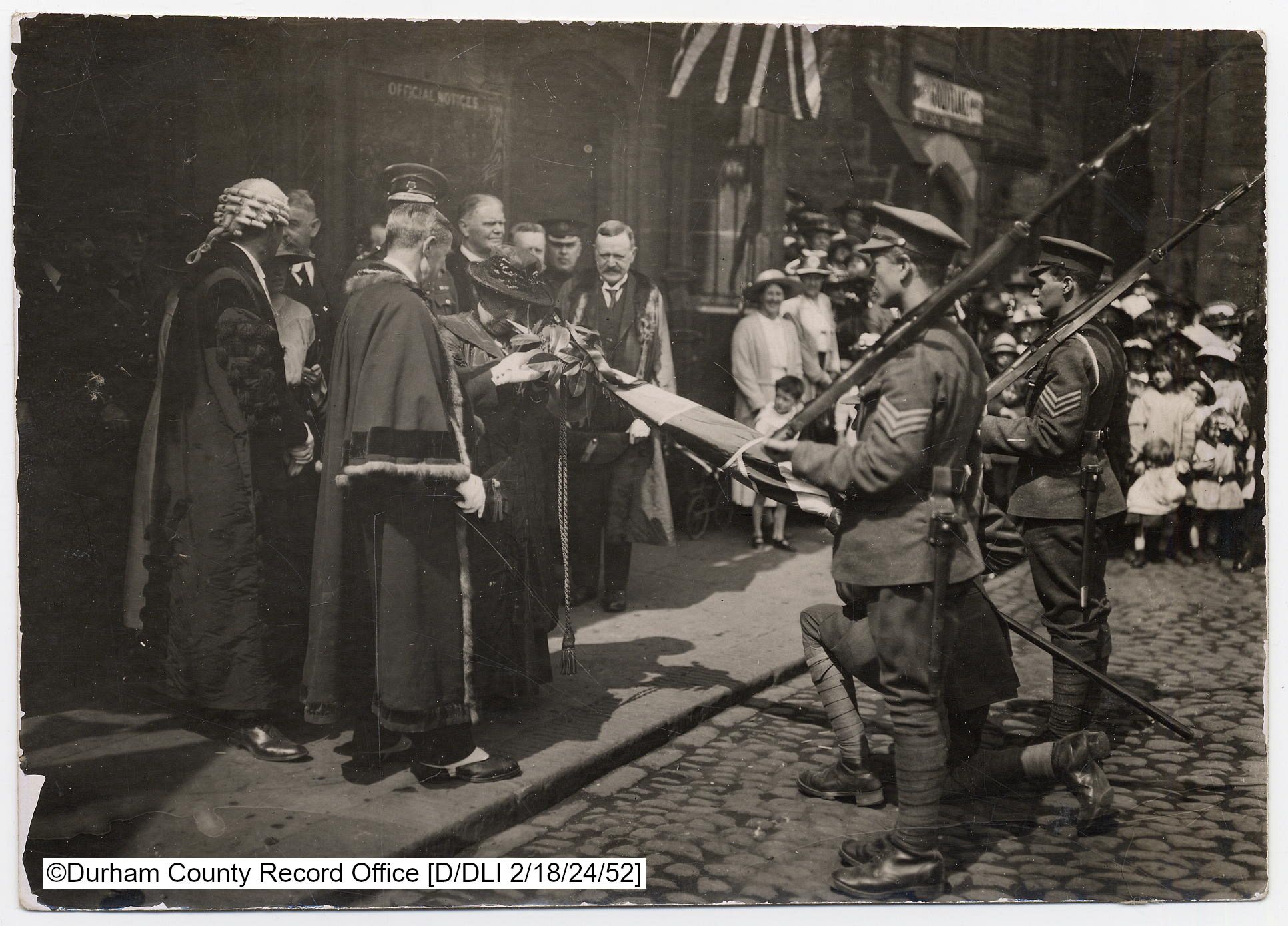 Black and white photo with a group of smartly dressed onlookers watching DLI soldiers present their Colours to local dignitaries in ceremonial clothing.