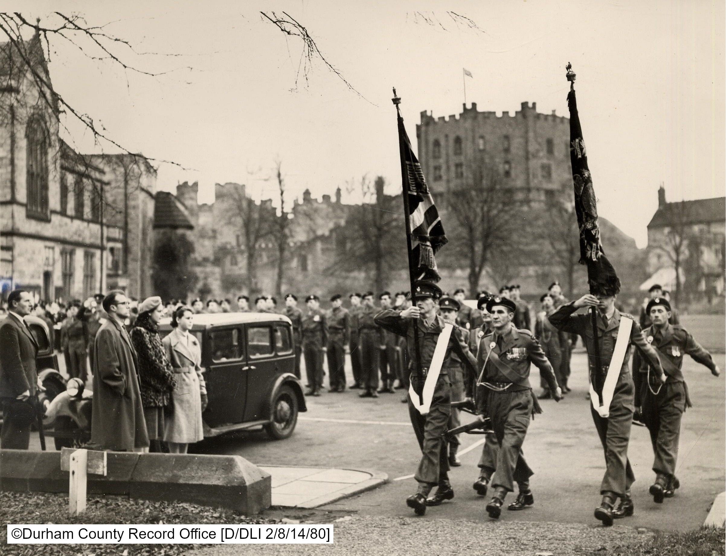 Black and white photo of DLI soldiers marching their Colours into Durham Cathedral to be laid up. A crowd of onlookers watch on from the left and Durham Castle can be seen in the background.