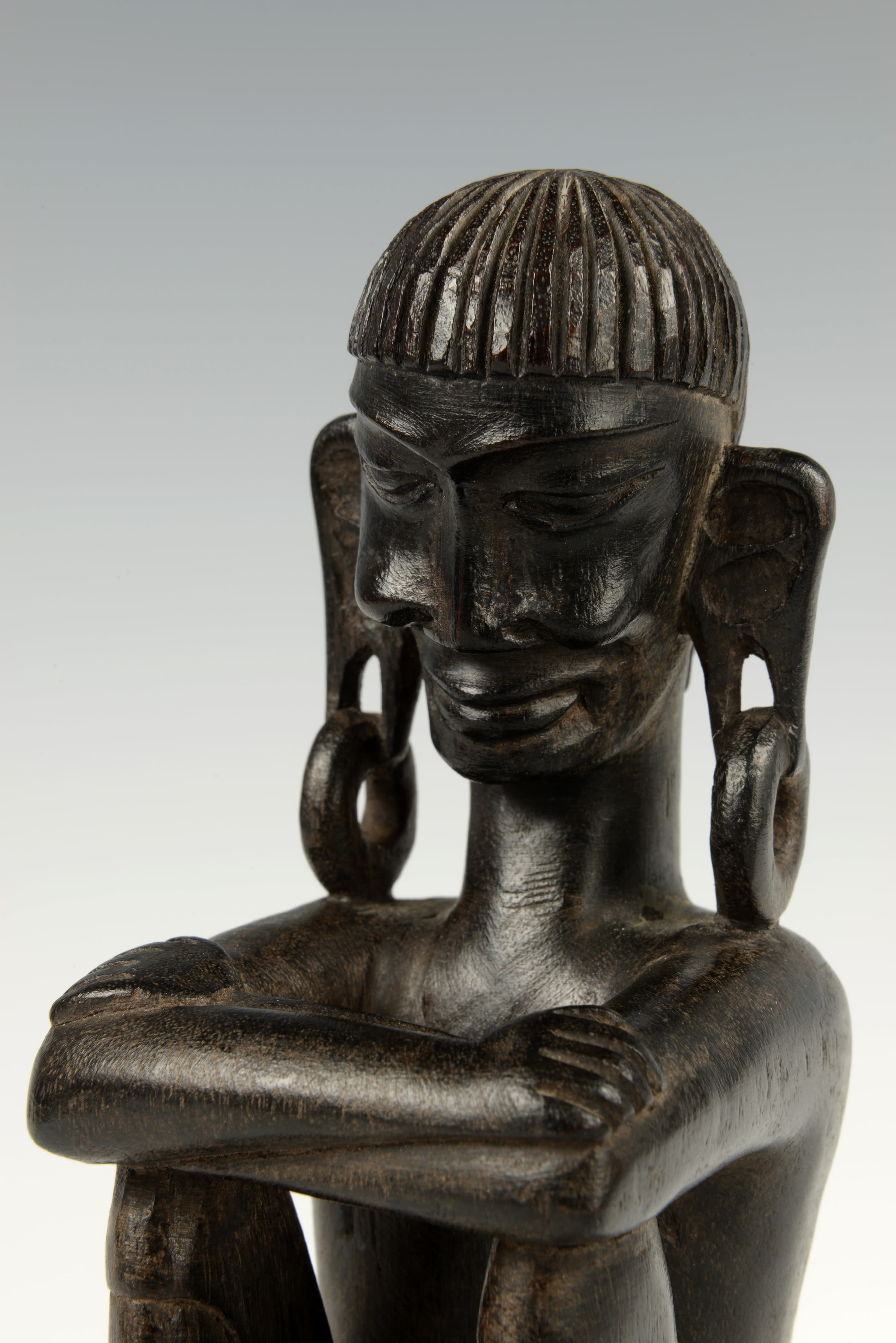 The man in this sculpture sits with arms folded on his raised knees. His ear lobes are dramatically pulled down by large hooped earrings.