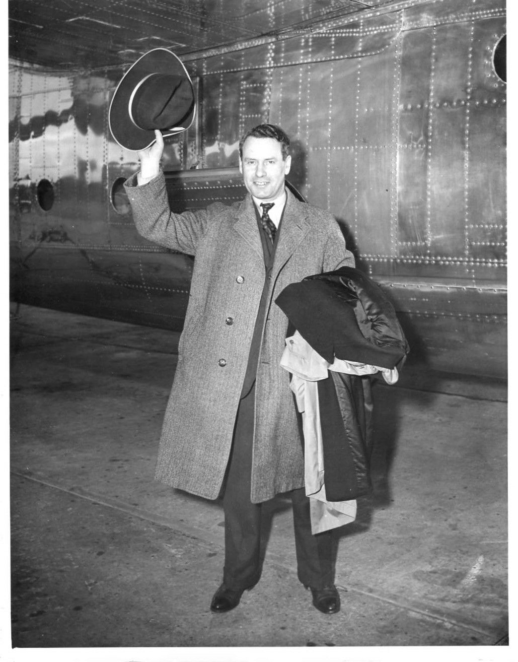 Malcolm MacDonald holding his coat in one arm, with hat held aloft in the other. He appears to stood next to a ship, waving either hello or goodbye.