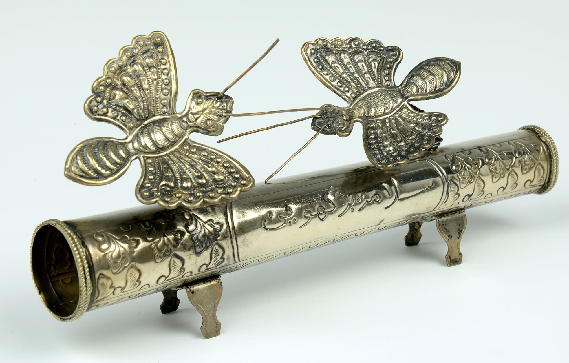 Long, cylindrical tube with patterned carvings. Above the tube two butterflies are fixed, with elaborately designed wings and tails.