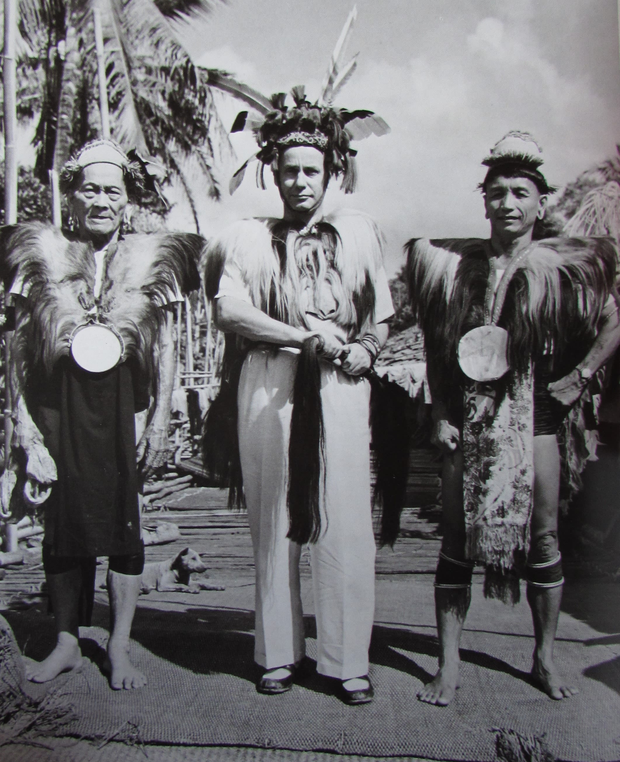 Malcolm MacDonald stands in the centre of two men, dressed in elaborate clothing with large hats and jackets made from animal furs and feathers.