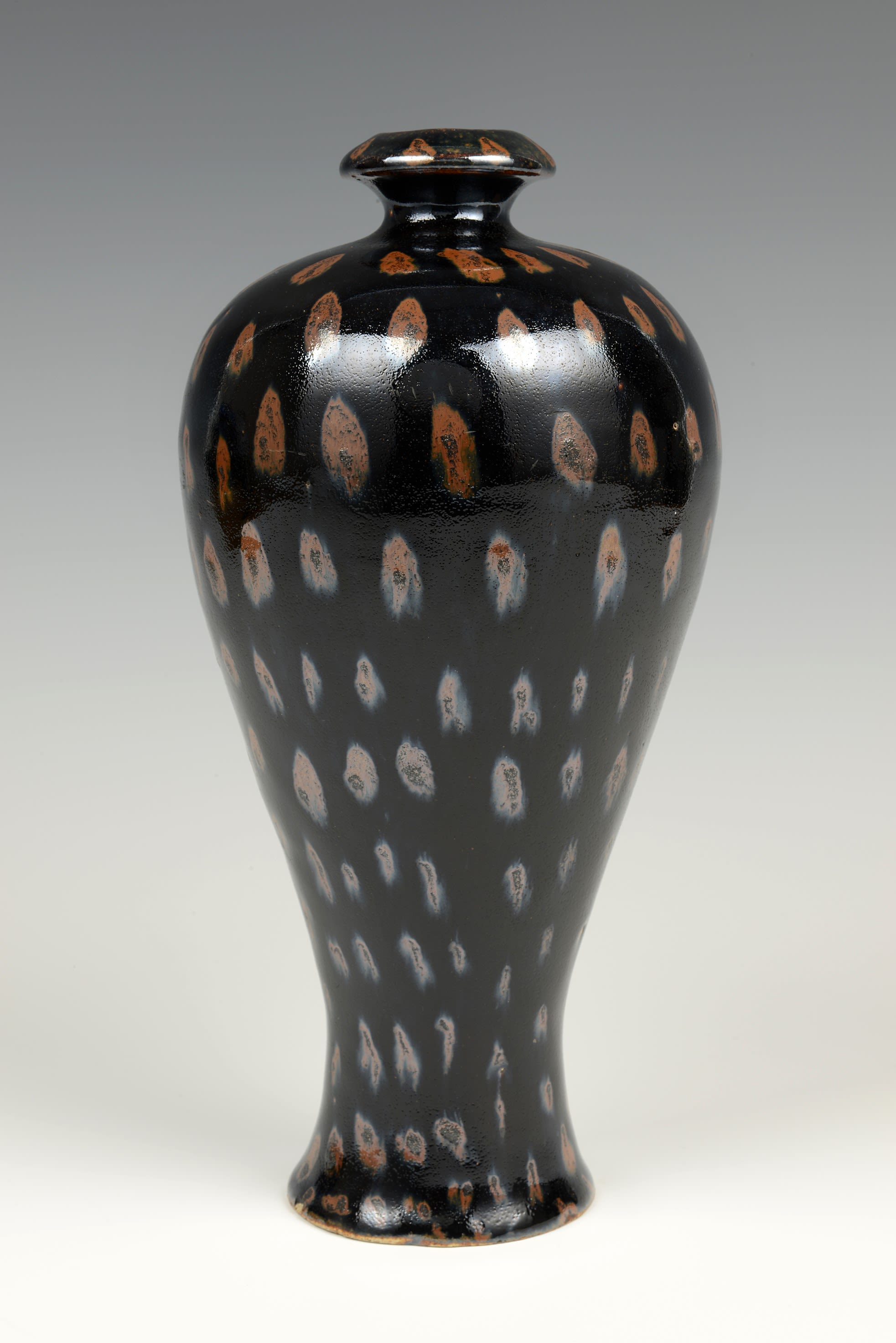 Tall black vase with oval shaped brown patterns running across its body. The vase has a narrow top, then grows wider, before thinning to a narrow base.