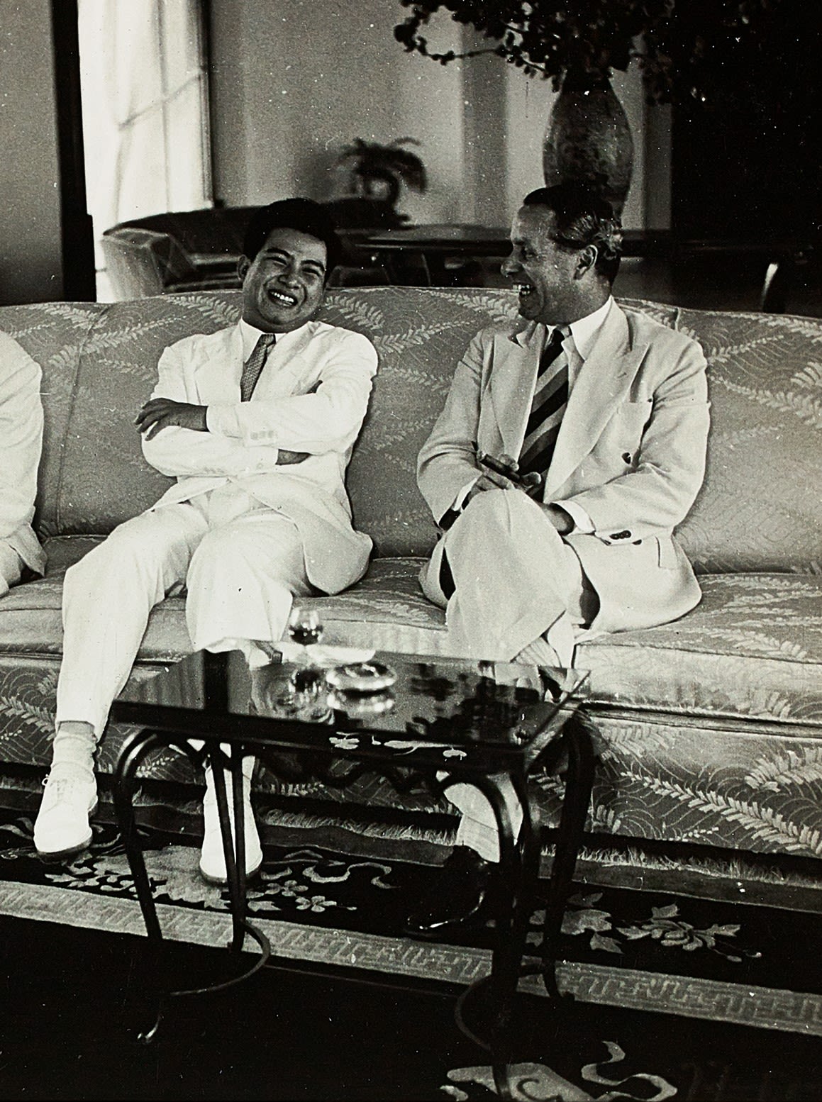 Malcolm MacDonald sits on a sofa with King Norodom Sihanouk of Cambodia. The two men are smiling and laughing, clearly enjoying a joke.