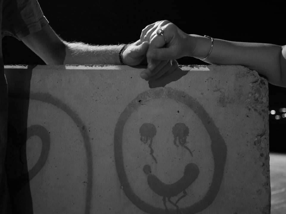 This black and white photograph taken by Shaho Omar is titled Concrete Love. It shows a smiley face painted onto a piece of concrete in Freedom Park. The paint runs slightly down the eyes, almost looking like tears. Two hands are clasped, resting on the top of the concrete. The concrete is the lightest part of the image.