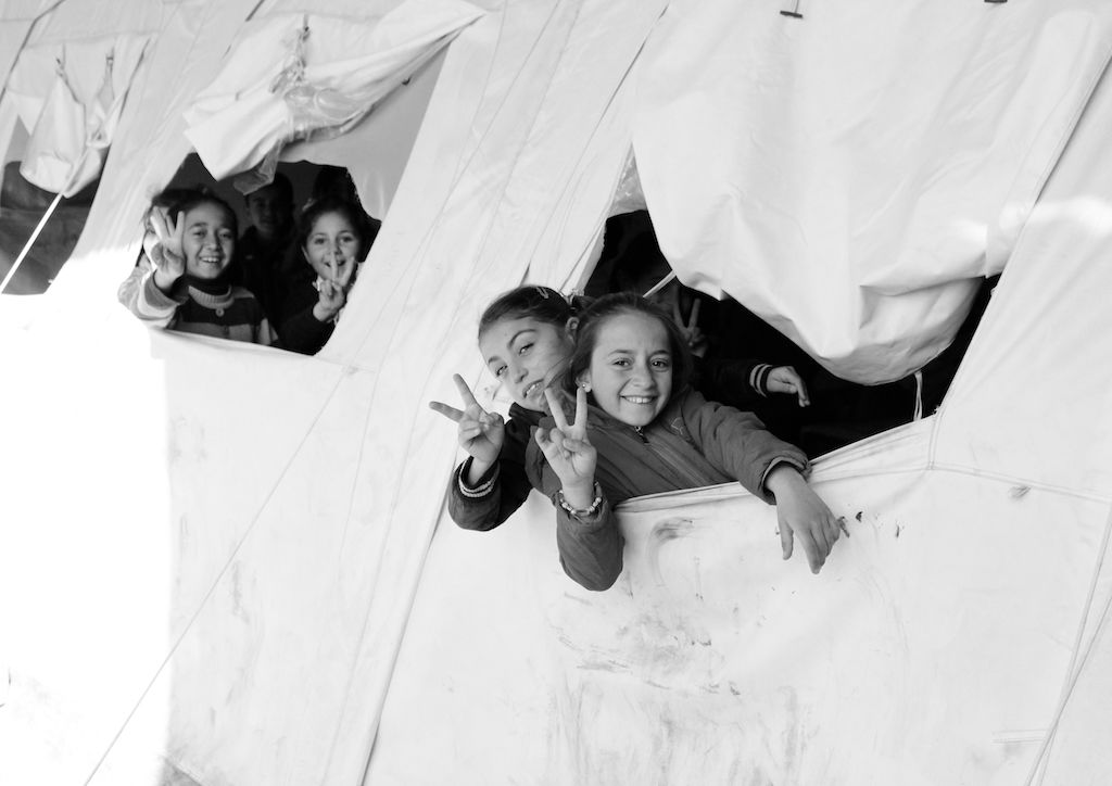 This black and white photograph taken by Ako Ismail is titled Victory. This is a very light image, the background is a white tent. From inside the tent, two groups of two children are peeking out of the tent and smiling at the camera. They have two fingers up in the victory symbol. 