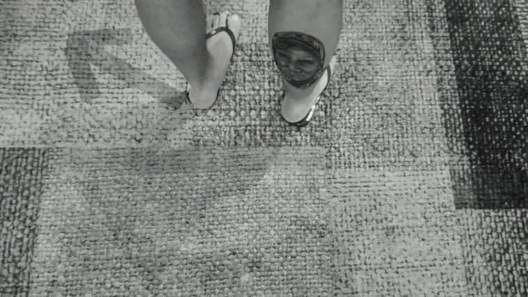 This black and white photograph taken by Shaho Omar is titled Mother. It shows the backs of a man’s calves where on the right hand side there is a tattoo of his mother’s face. The background is of the floor which is a patterned grey with the shadow of a happy face. The man’s sandaled feet are stood over where the nose and eyes of the face on the floor would be. Next to the left leg is an arrow pointing diagonally North-West.