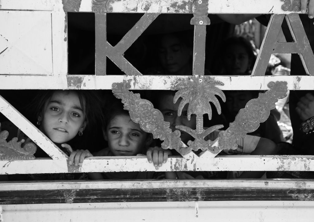 This black and white photograph taken by Ako Ismail is titled Search for Safety. In the foreground of the image are three children who are peering out from behind some panelling. In the background are some other children who cannot be seen very clearly because of the light levels. The child on the right is obscured quite a lot by the panelling whereas the child in the middle is looking out, away from the camera, and the child on the left is looking directly at the camera. All three have worried expressions on their faces. 