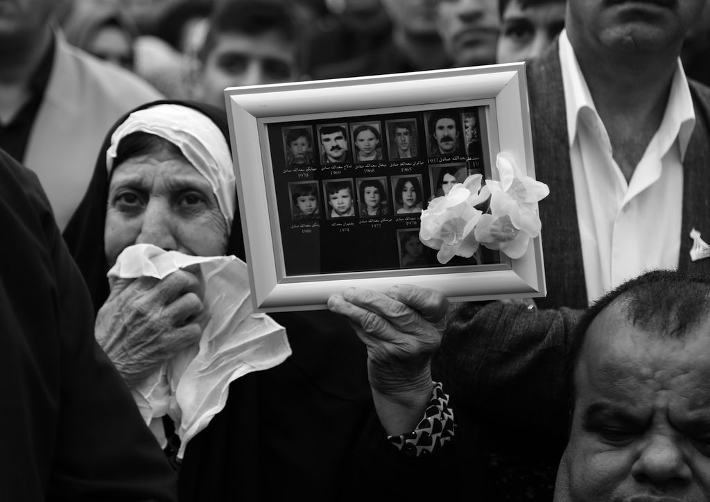 This black and white photograph taken by Ako Ismail is titled All were Lost. It shows a woman wearing a head covering, holding a handkerchief up to her mouth with a grieved expression on her face. In her left hand she is holding up a frame in front of the camera lens showing photographs of her eleven family members who were all killed in the chemical attack. Other people can be seen in the background although they are partially hidden by her and the frame. 