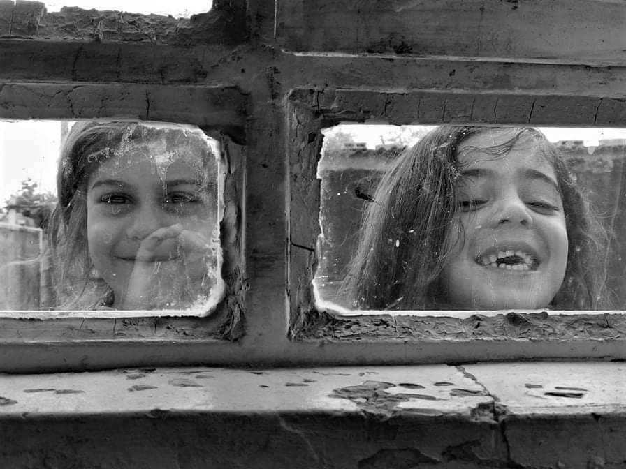 This black and white photograph taken by Shaho Omar is titled Reflections. It shows two young girls who are smiling directly at the camera. They are stood behind a window and are framed by the frames of the window. 