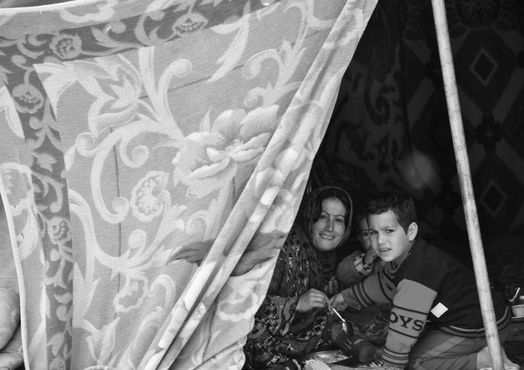 This black and white photograph taken by Ako Ismail is titled Family Life. It shows a woman and young boy on the right hand side of the photograph, smiling at the camera lens. There is a light, patterned tent that is opened so that we can see these people.