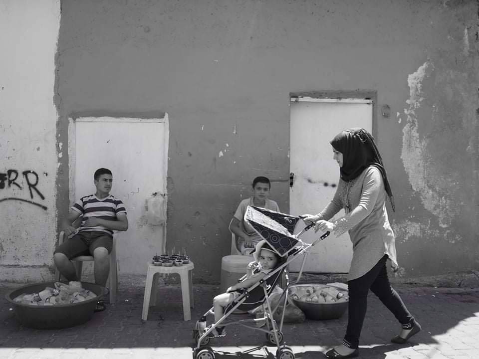 This black and white photograph taken by Shaho Omar is titled The Water Sellers. It shows a young man and a boy sat on stools in front of a building that has two doors. The boy on the right hand side is looking directly at the camera whilst the young man is looking towards the boy. They are both wearing shorts and t-shirts. Between them there is a table where they are selling their water as well as two baskets on the floor. There is graffiti on the building in the background. To the right hand side of the image is a person pushing a pushchair with a small child in it who is also looking at the camera. 