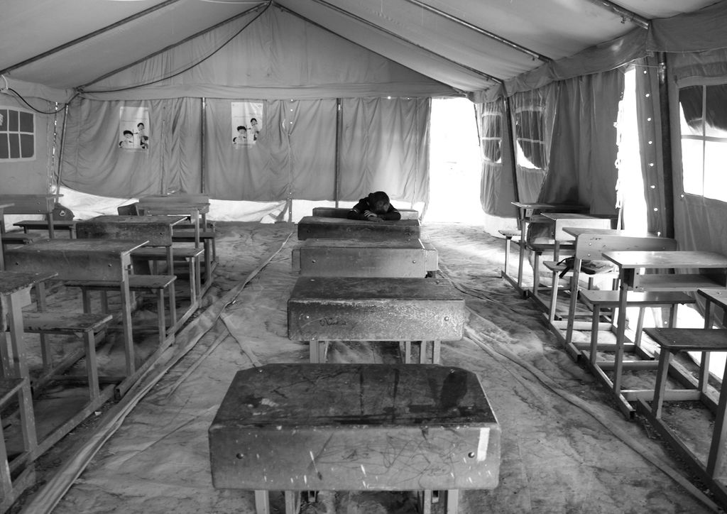 This black and white photograph taken by Ako Ismail is titled Back to School. It shows a classroom which is inside a large tent and has a temporary floor. There is light coming in from an opening in the tent at the back of the classroom. It is filled with desks, all of which are empty apart from one in the middle of the back row where there is a child with their head on their arms on the desk. There are no other people in the image. 