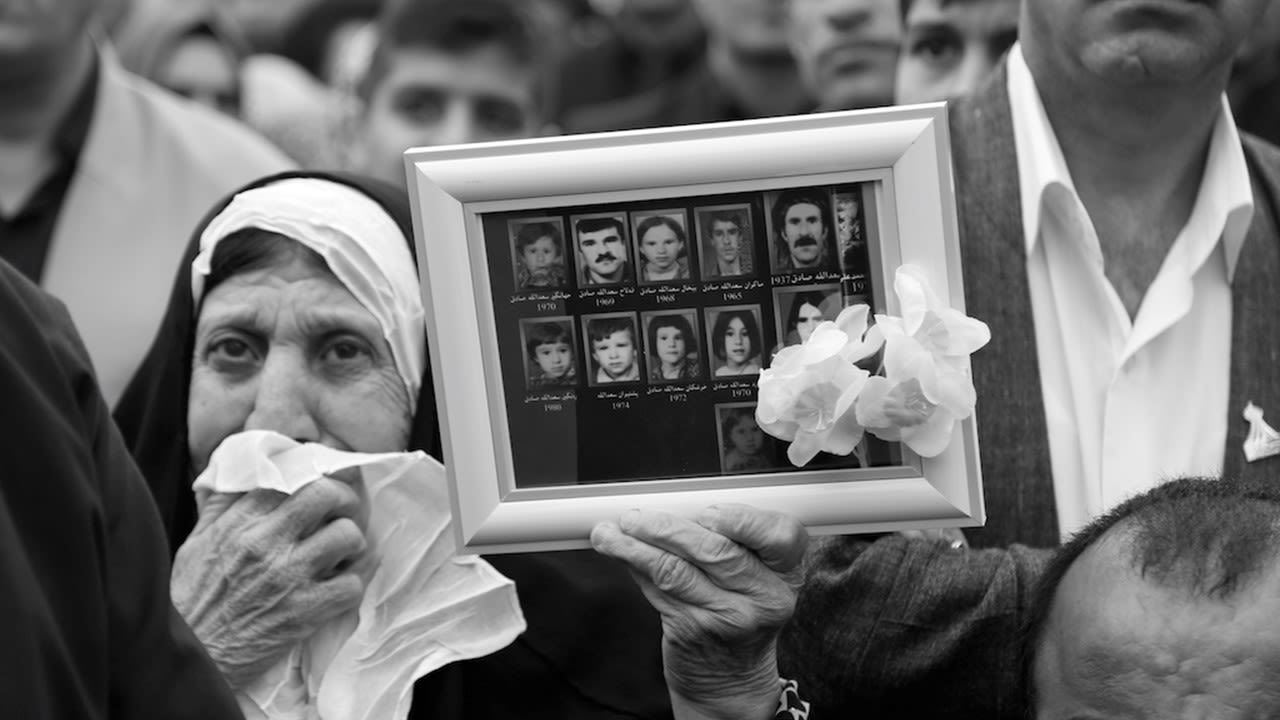 This black and white photograph taken by Ako Ismail is titled All Were Lost. It shows a woman wearing a head covering, holding a handkerchief up to her mouth with a grieved expression on her face. In her left hand she is holding up a frame in front of the camera lens showing photographs of her eleven family members who were all killed in the chemical attack. Other people can be seen in the background although they are partially hidden by her and the frame. 