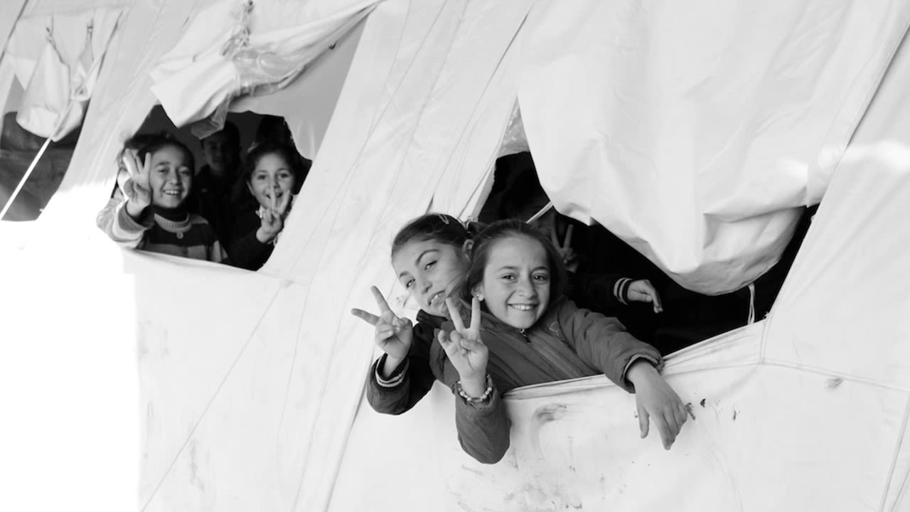 This black and white photograph taken by Ako Ismail is titled Victory. This is a very light image, the background is a white tent. From inside the tent, two groups of two children are peeking out of the tent and smiling at the camera. They have two fingers up in the victory symbol. 