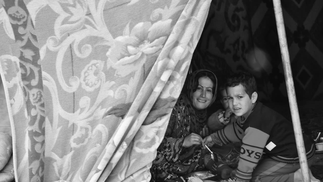 This black and white photograph taken by Ako Ismail is titled Family Life. It shows a woman and young boy on the right hand side of the photograph, smiling at the camera lens. There is a light, patterned tent that is opened so that we can see these people.