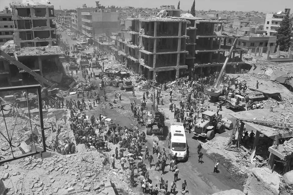 This black and white photograph taken by Shaho Omar is titled Suicide Attack. It shows the aftermath of a suicide track bomb that hit multiple Kurdish security buildings in Qamishli in Syria, injuring more than 100 people and killing 40. The image shows buildings standing in the background with the foreground showing lots of people on the ground as well as some vehicles. The picture is quite zoomed out so individual people are not identifiable and the scale of the buildings to the people can be seen.