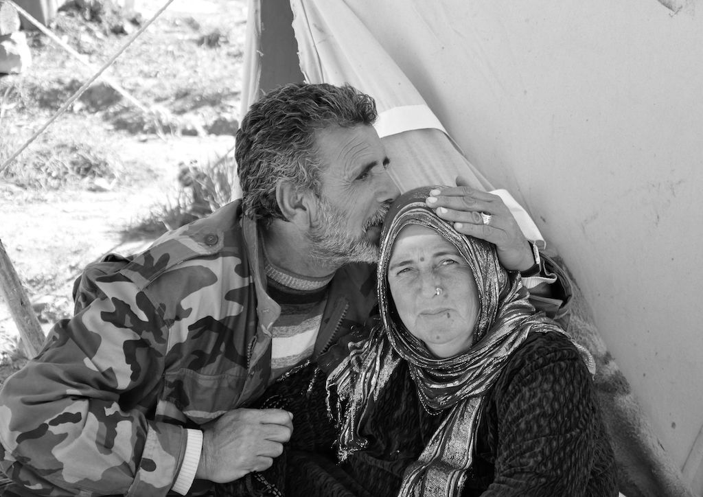 This black and white photograph taken by Ako Ismail is titled The Kiss. It shows a man on the left who is kissing the side of a woman’s head. He has his left hand on her head as he does this, so that you can see he is wearing a ring on his ring finger, and a watch on his wrist. He is looking to the right of the image, over her head whereas she is looking directly at the camera lens. She is faintly smiling and has both a nose ring and a head covering on. 