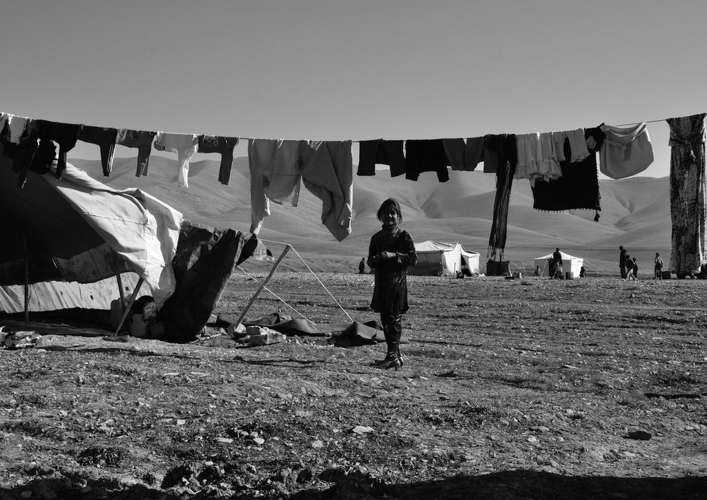 This black and white photograph taken by Ako Ismail is titled A Different Daily Life. It shows a girl in the centre of the foreground who is smiling at the camera. Behind her is a line with clothes hung up on it and in the left hand side of the image is a tent. Further away in the image are other tents and the figures of other people.
