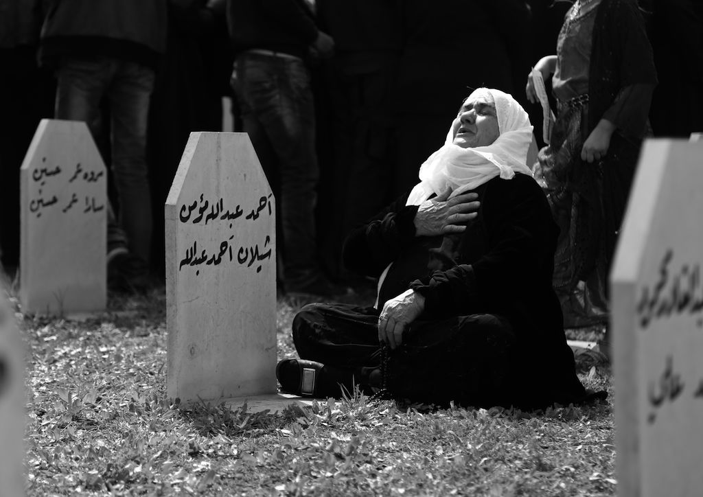 This black and white photograph taken by Ako Ismail is titled A Mother’s Cry. It shows a woman sat cross-legged on the floor, alone. Her hand is on her chest in anguish with a pained expression on her face. Her head covering is white and doesn’t cover her face, whilst the rest of her clothes are all black. There are three graves in the image as well as three people in the background whose heads cannot be seen in the frame.
