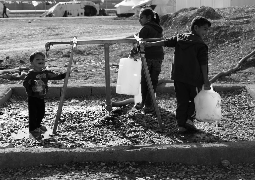 This black and white photograph taken by Ako Ismail is titled Meaning of Life. It shows three young children at a water pump. The two taller children are both holding a water cannister each. No other structures can be seen in the photograph, showing how these pumps can be far away from some camps, meaning the children have to carry these heavy containers on their walks back.