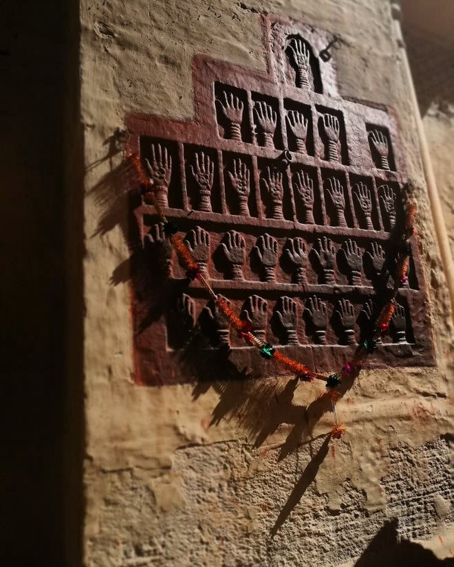 A close up photograph of a wall carving. Showing over 30 small palms of hands with a necklace pin over the top.