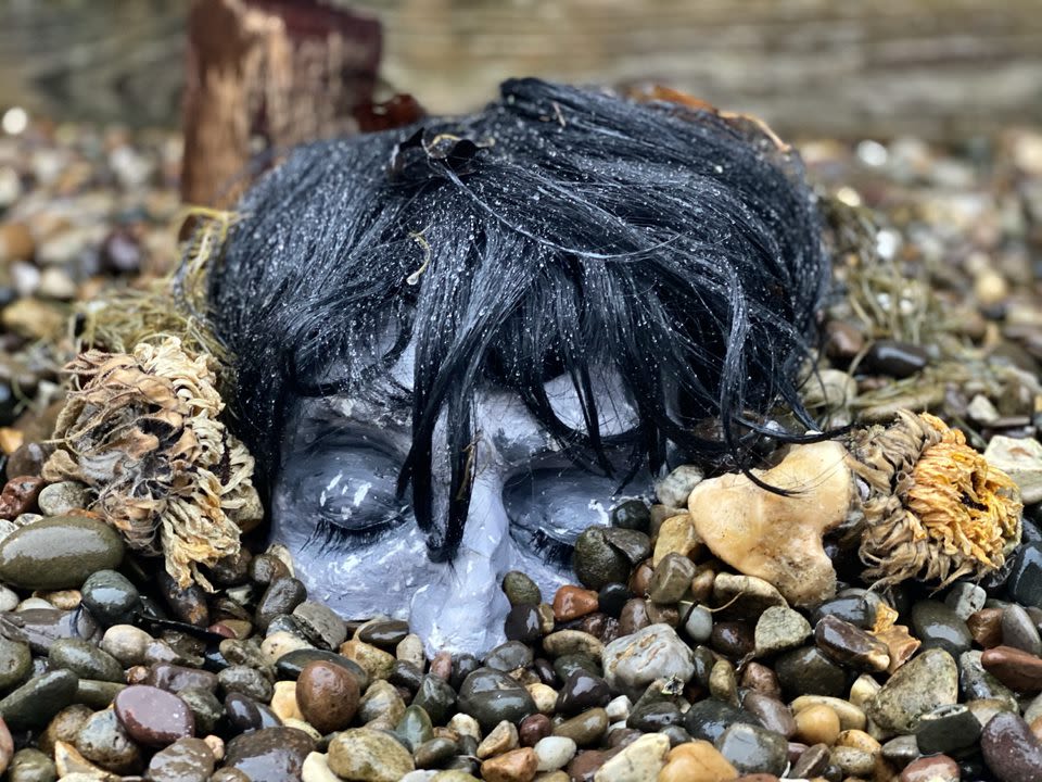 Photo of an installation. A plaster head buried in stones, showing only the head from the nose up, with short black hair. Surrounded by a dead flower, bone and moss.