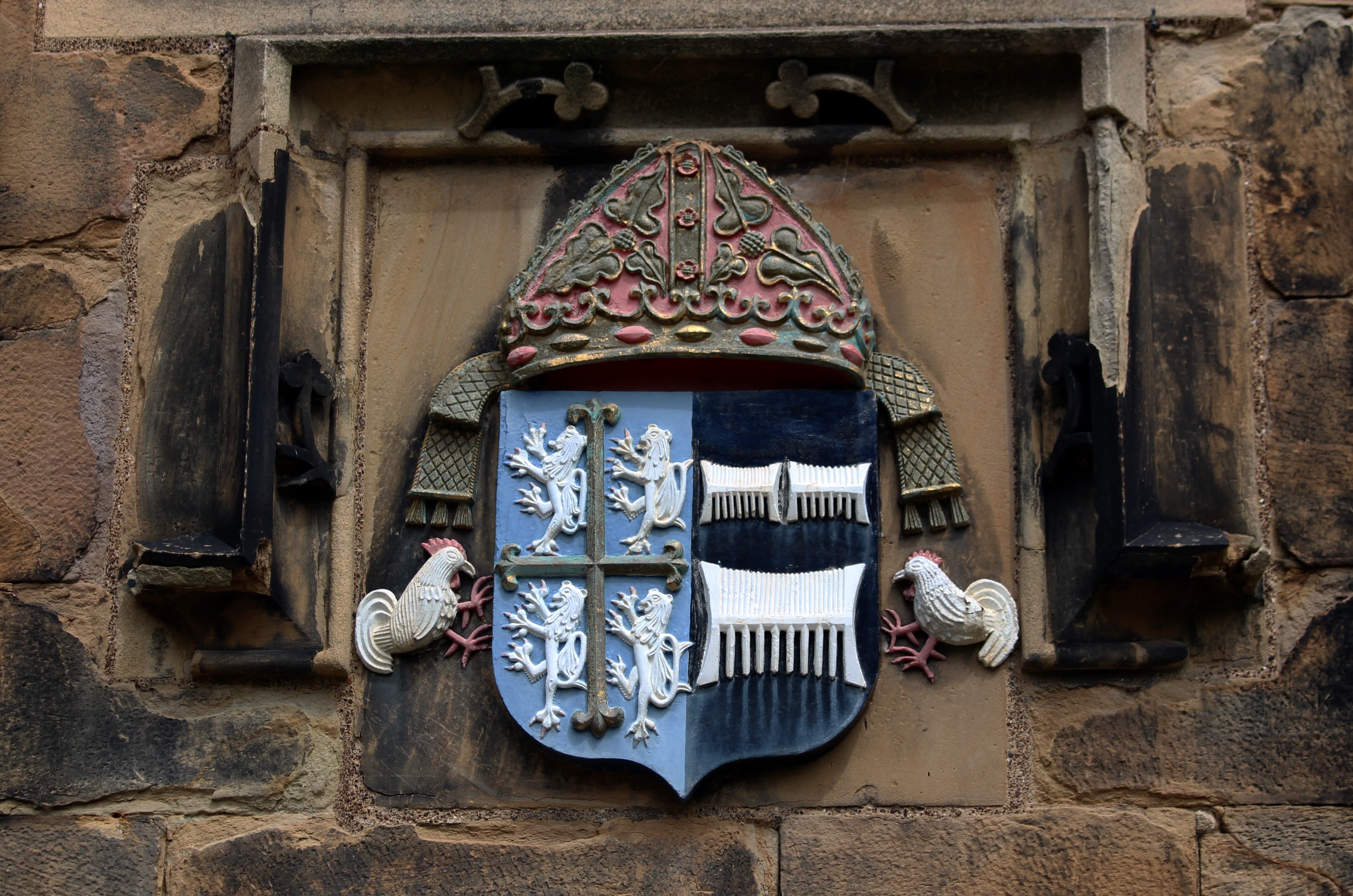 Tunstall family coat of arms, as seen on the courtyard wall of Durham Castle. The design of the shield includes roosters, lions and combs. The shield is topped with the ceremonial mitre of the Prince Bishops.