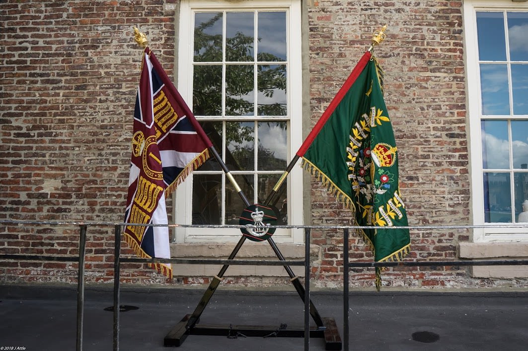 Photograph of DLI King's and Regimental Colours, displayed outside on crossed poles. The regimental colour has a green background, the King's Colour is a Union Flag with a golden trim.